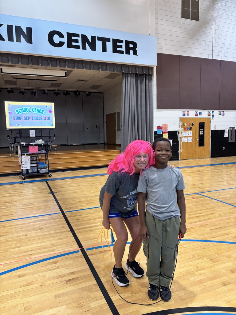 Two individuals pose in a gymnasium with a "Skin Center" sign in the background. A person with pink hair smiles at the camera.