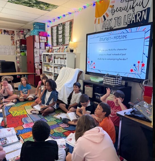 A group of children in a classroom, some sitting on the floor with colorful mats, focused on a large screen displaying text.