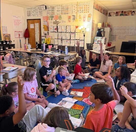 A classroom with a teacher leading a discussion. Students sit on the floor with papers and books.