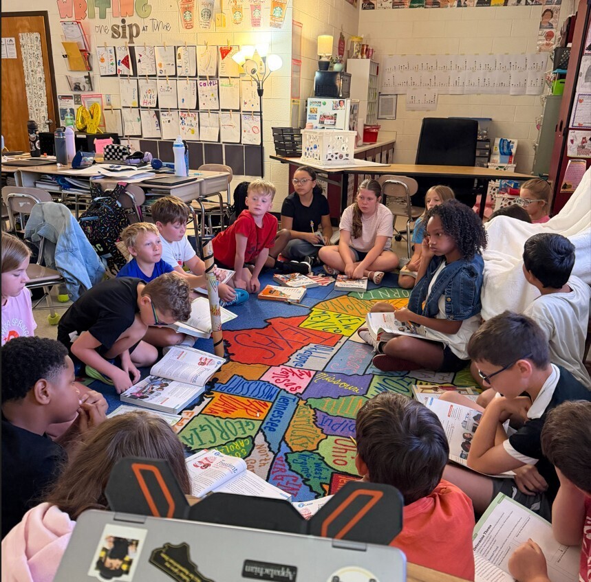 Classroom with children sitting in a circle around a colorful map, reading books and papers.