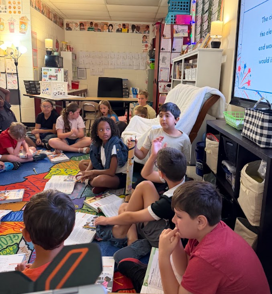 Children sit in a circle on the floor, facing a screen. Some hold books and papers. Bright colors on the floor.