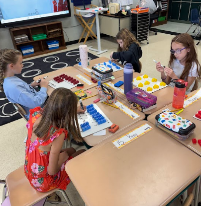 Four children seated at desks with educational materials. One has a colorful placemat.