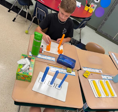 A child seated at a desk works with colored rods and paper, using a tissue box and a box of crayons.