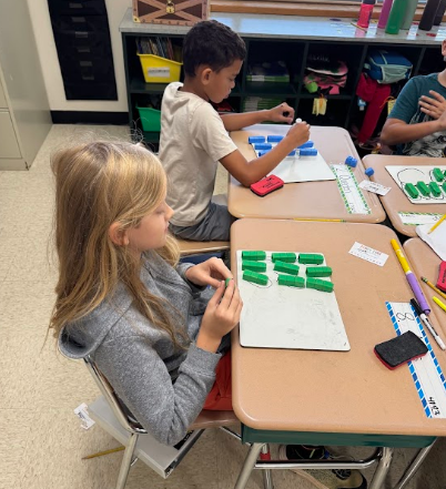 Students seated at desks, using materials. One student works with green objects, while another has a red object.