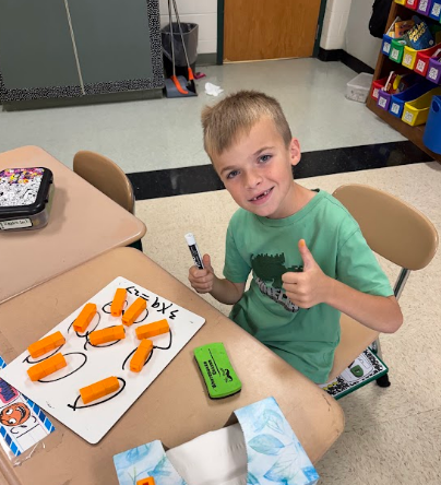 A young boy sits at a table, smiling and giving a thumbs up. He holds a marker.