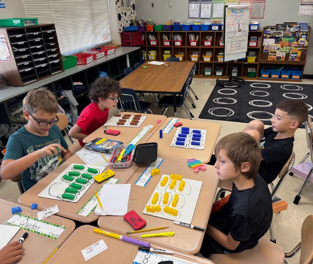 Four boys seated at a table in a classroom. Each child uses a different set of colored manipulatives.