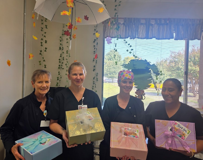 Four women in dark uniforms hold gift boxes. Behind them is a window with a view of trees.