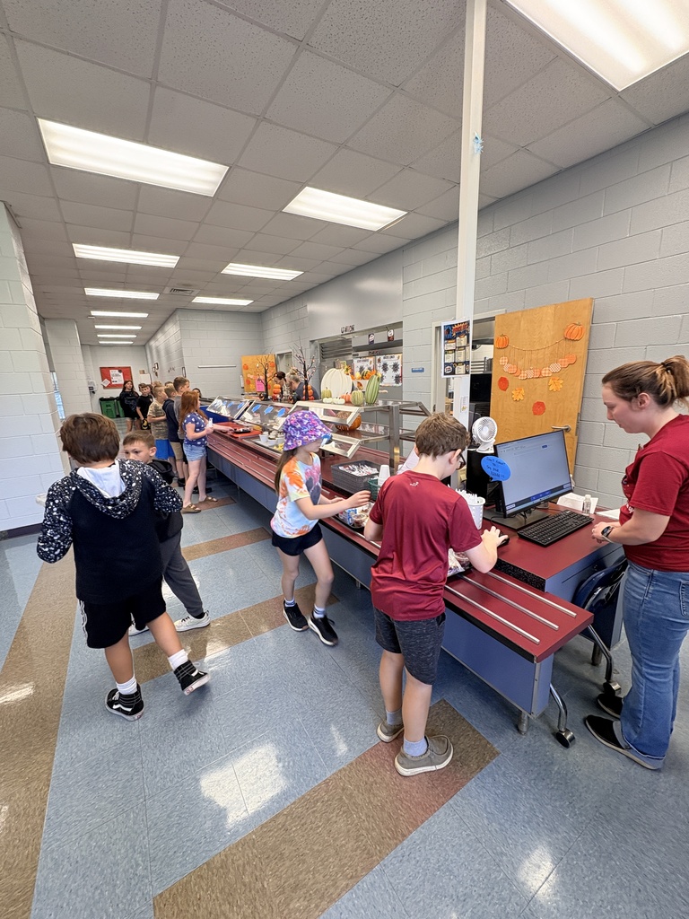 Kids waiting in line at a cafeteria counter with an adult and monitors on the table.