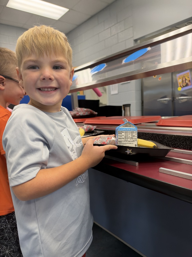 Boy smiling at cafeteria table with tray of food, bottle, and another child behind.