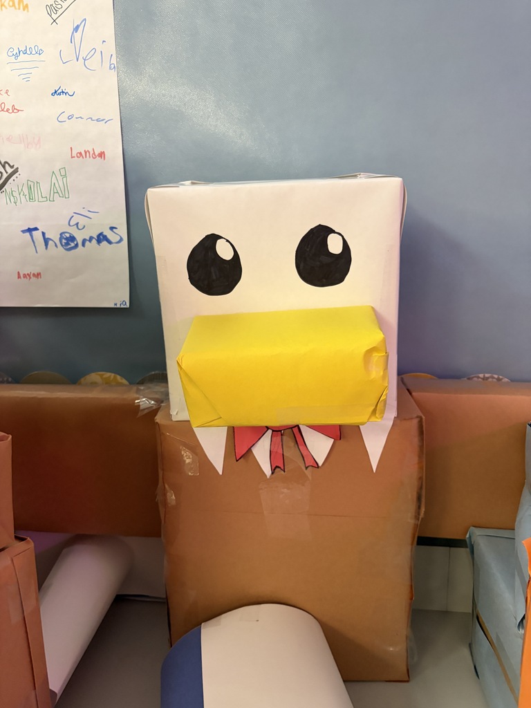 A brown box with a yellow beak and black eyes stands on a table near a whiteboard.