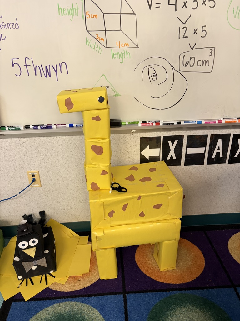 A yellow object resembling a giraffe stands on a multicolored floor. Behind it is a whiteboard with diagrams and text.
