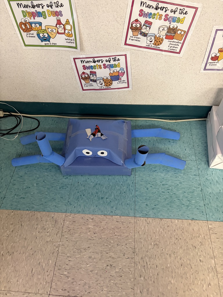 A blue toy with eyes and a face sits on a blue carpet. Behind it, a wall has four colorful posters.