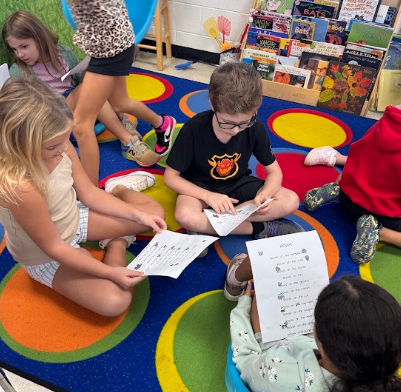 A group of young children in a classroom setting, sitting on a multicolored rug, holding papers.