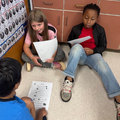 Three children sit on the floor reading papers. One holds a sheet with printed text. Behind them is a bulletin board with images of faces.