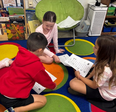 Children sit on a colorful rug, reading papers. One girl sits in a chair behind them.