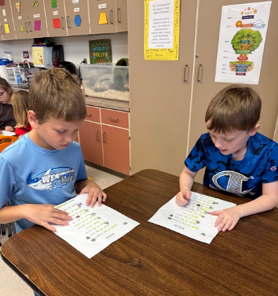 Two boys seated at a table, each holding a paper with green text, focusing on their work.