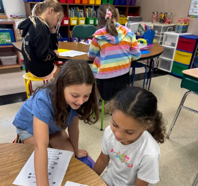 Two students work together at a table in a classroom with shelves of books in the background.