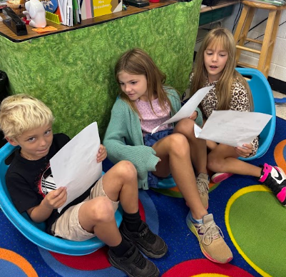 Three children seated in blue chairs on a colorful rug, holding papers, reading or discussing.