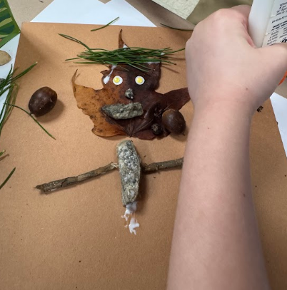 A student works on a project using acorns, leaves, and twigs on a brown background.
