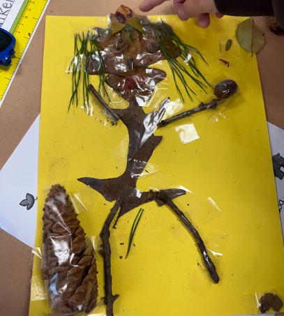 A student's artwork on yellow paper. The piece is a human figure made from twigs, leaves, and a pine cone.