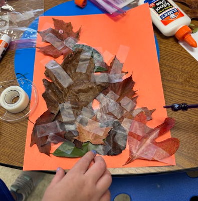 Hands place leaves on orange paper. A table has paper, glue, tape, scissors, and plastic.