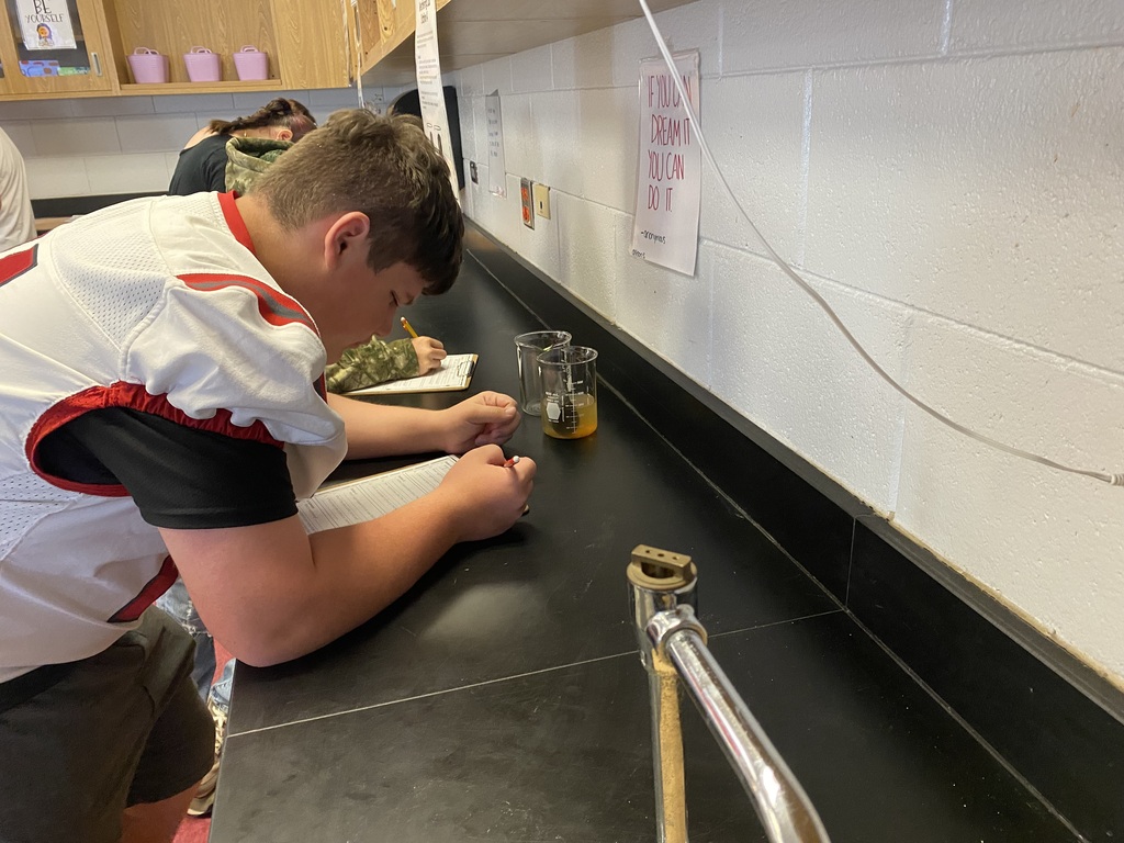 A student sits at a lab counter, writing in a notebook and working with a small yellow liquid.