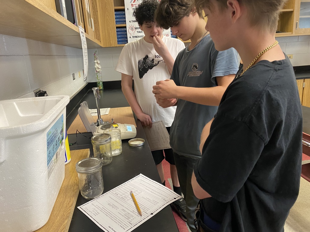 Three students examine a table with a jar and notebook, while one holds a pencil.
