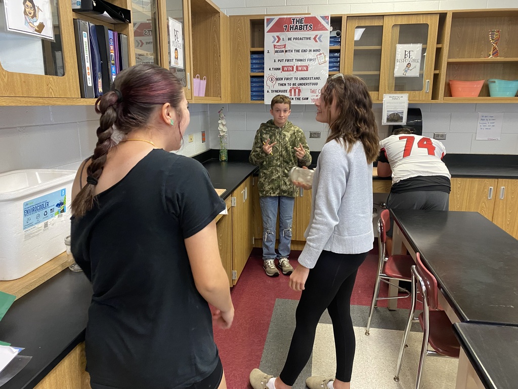 Students in a science lab. One student wearing a lab coat stands near a table. Two other students talk.