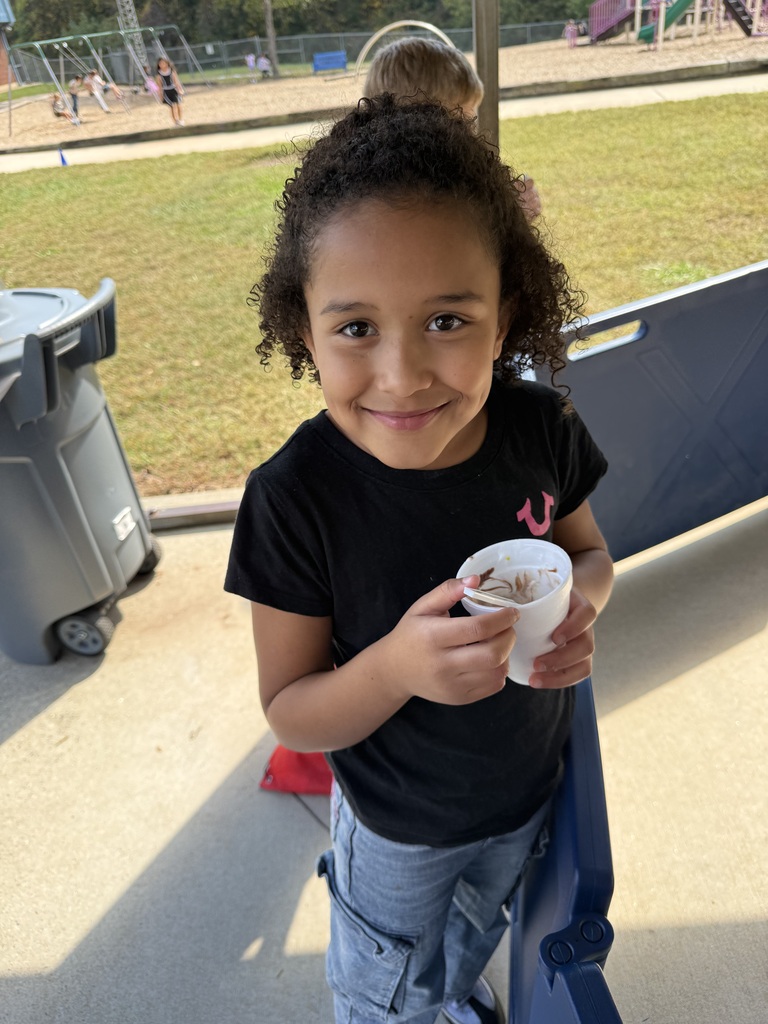 A young girl with curly hair stands outside holding a cup of ice cream. She smiles.