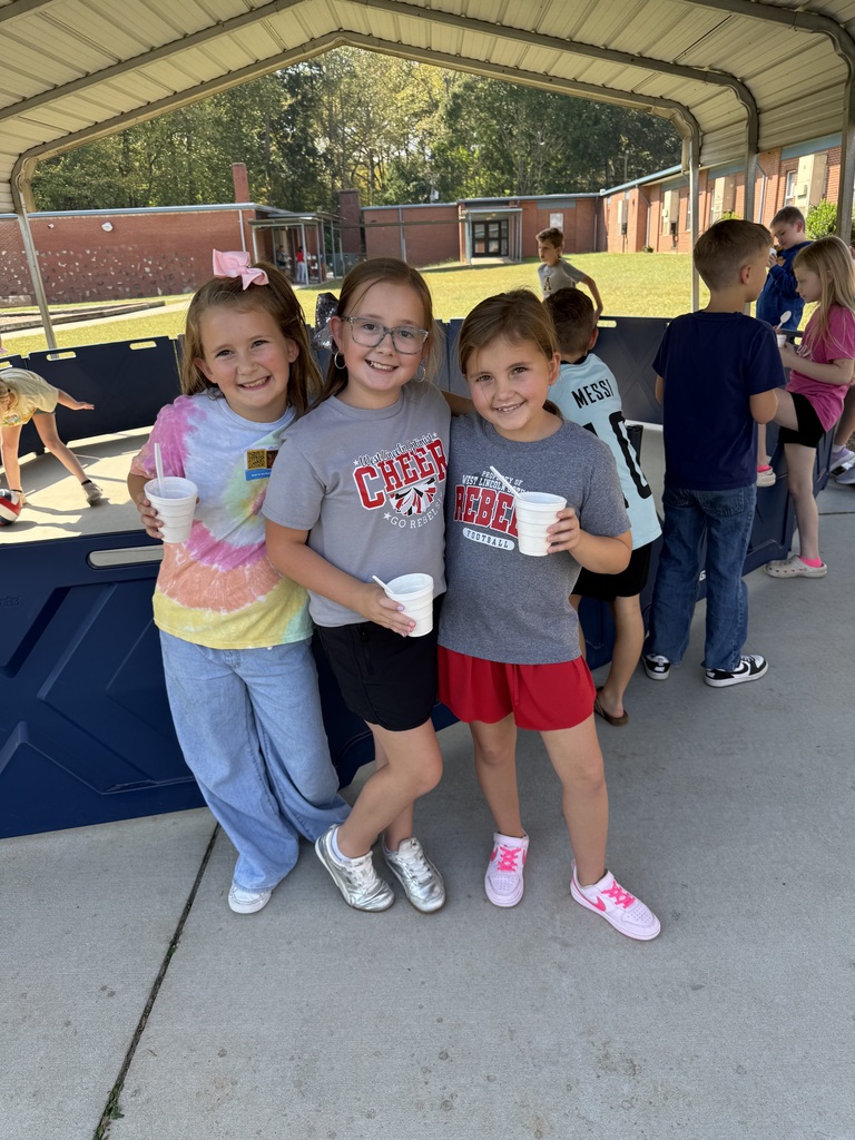 Three girls holding cups stand in front of a building with a canopy. Other children and adults are in the background.