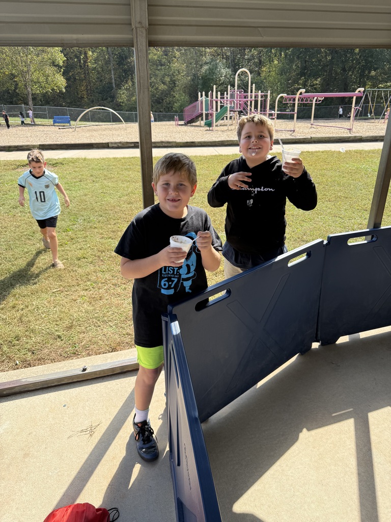 Two kids in front of a shelter, holding cups. A third child runs in the distance.