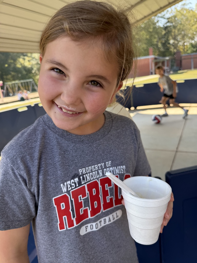 Child holds cup with spoon. Person in background runs towards soccer ball. Trees and fence in the background.