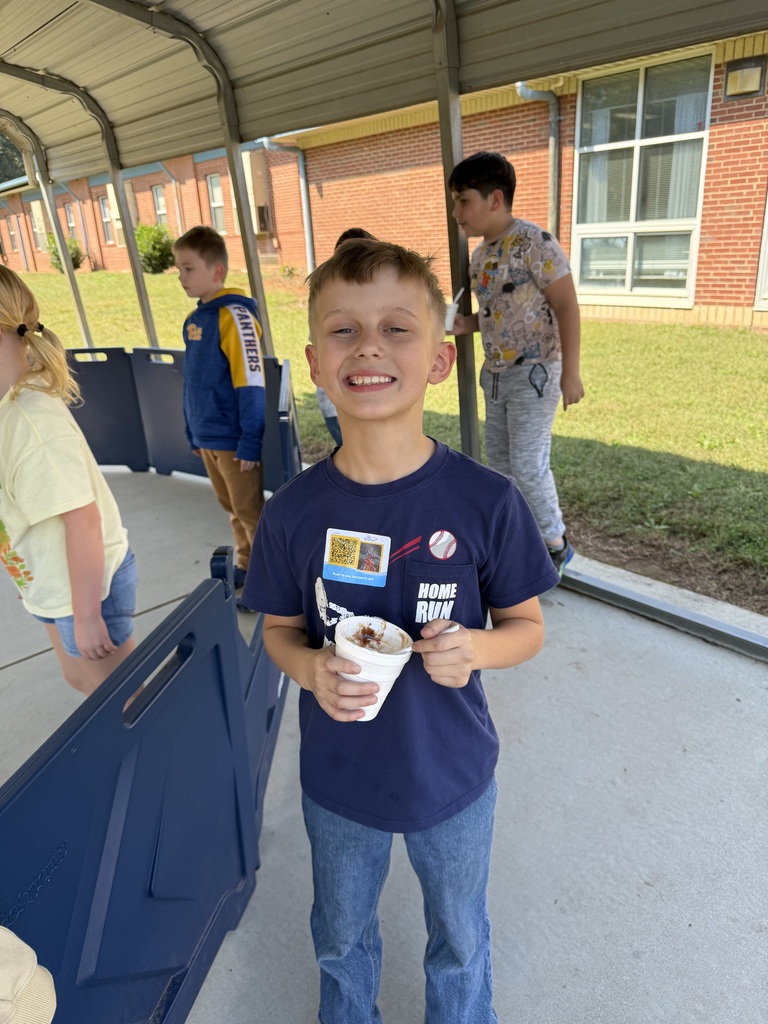 A smiling boy holding an ice cream cup stands under a covered walkway near a school building.