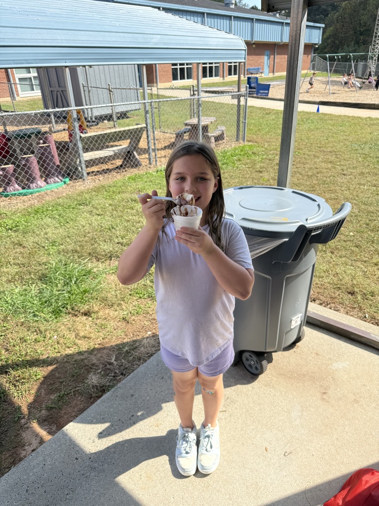 A young girl eats ice cream from a cup while standing on a patio by a trash can.