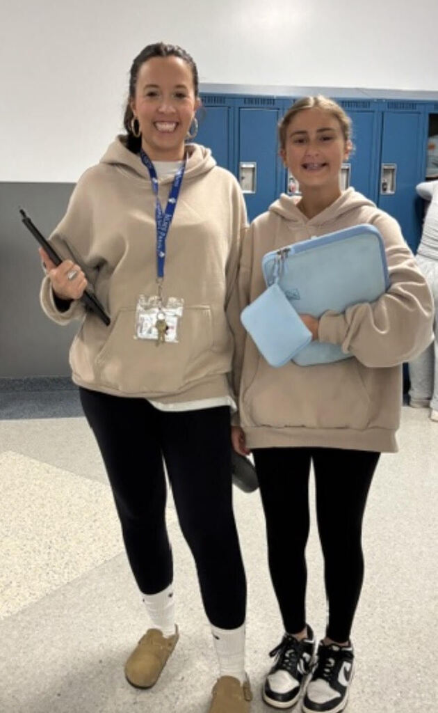 Two people stand in a hallway, both wearing beige hoodies, black pants, and sneakers. They hold items in their hands. Lockers are visible in the background.