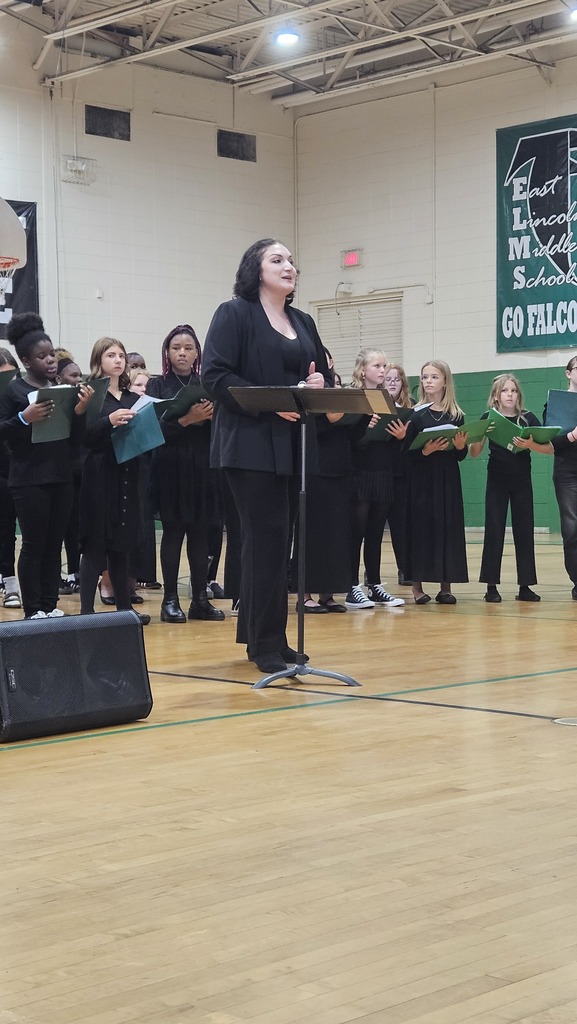 Woman standing at podium with microphone, addressing choir holding music stands on gym floor.