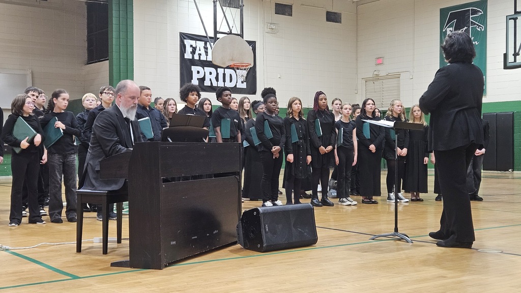 Choir performs with piano in a gymnasium; one person directs, others stand in rows, a banner reads "FAI PRIDE".