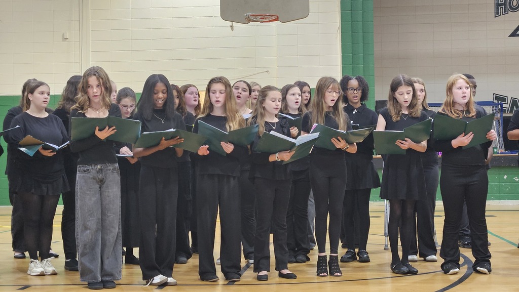 Choir members stand in a row on a basketball court, holding green songbooks and singing.