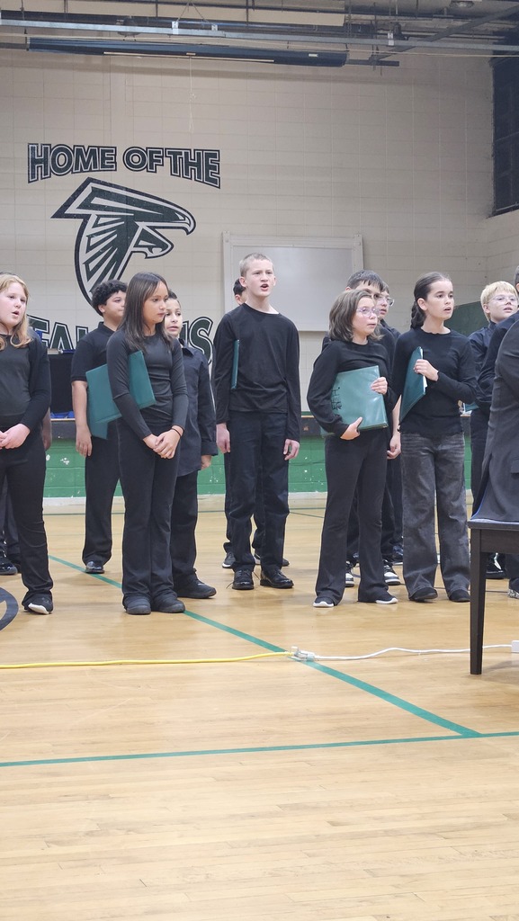 A group of students stands on a basketball court holding folders. A teacher stands at a piano.