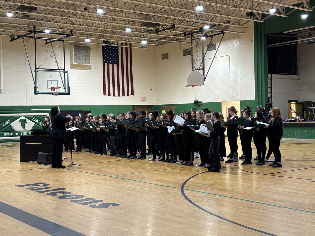 Choir in a gym, with a flag on a wall, a basketball hoop, and a speaker.