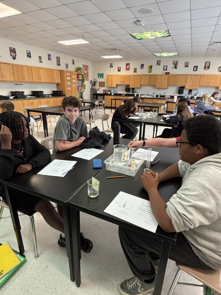 Four students in a classroom sit at tables. Three have papers and pens. One student holds a clear glass cup.