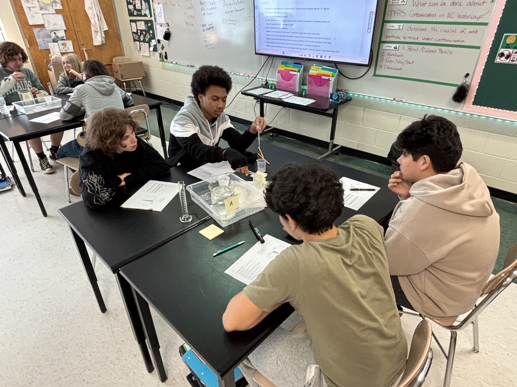 Students in a classroom setting working in groups at tables, with a projector screen and whiteboard visible.