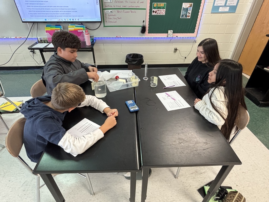 Four students sit at a table with a science experiment and papers. A screen is mounted on the wall behind them.