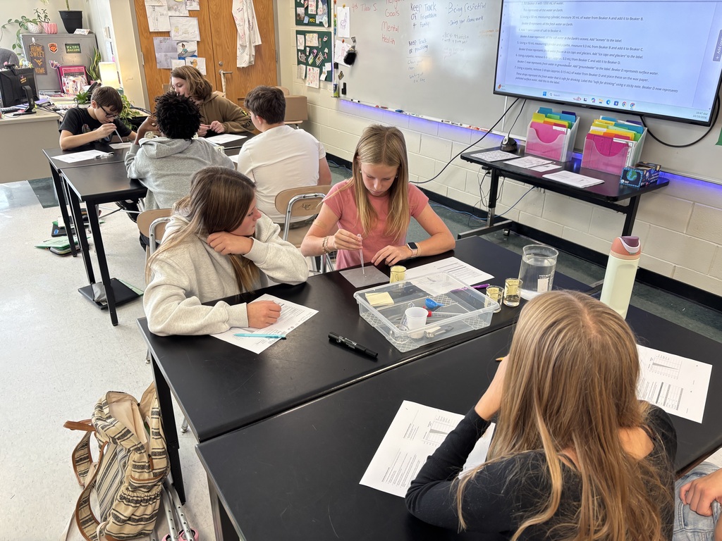 Students work at tables with papers and containers, facing a projector screen displaying text.