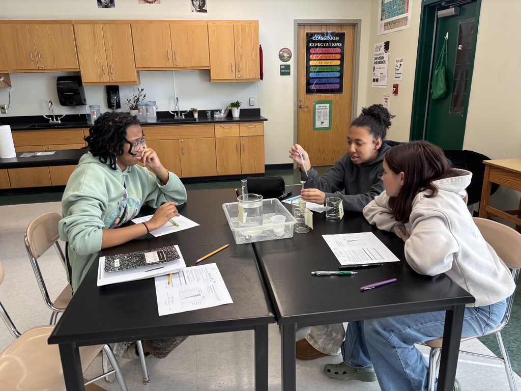 Four students seated around a black table with papers, pencils, and a tray of liquids.