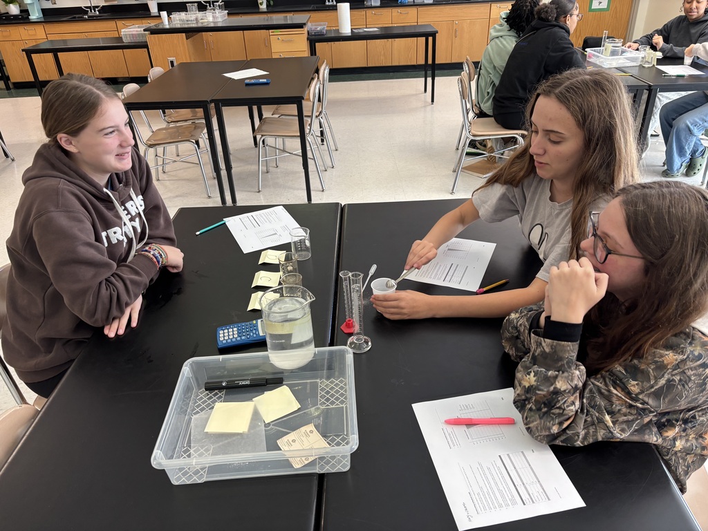 Three students work at a table with papers, a bottle of water, a pen, and a calculator.