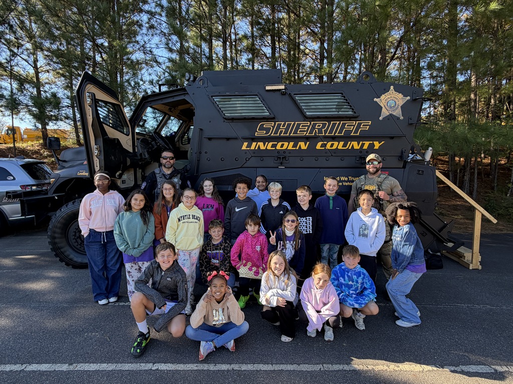 Children in casual clothing pose for photo in front of police vehicle in a parking lot.