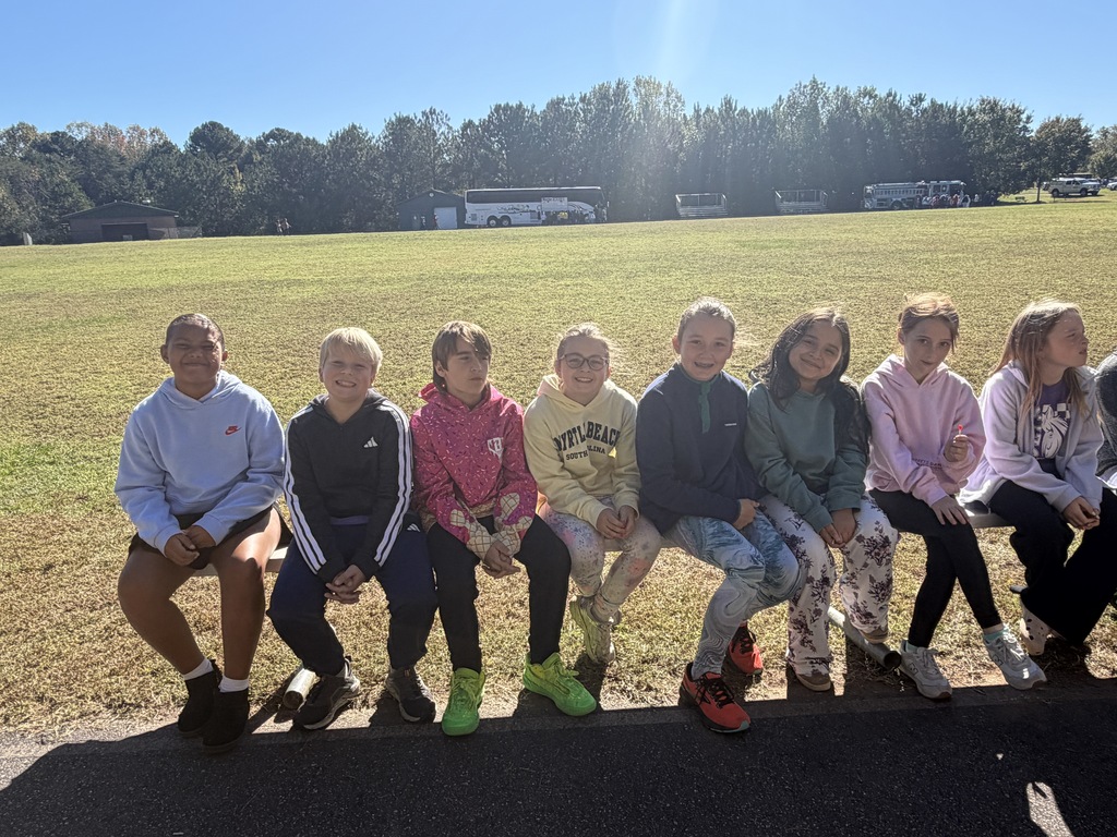 Nine children, wearing hoodies and sneakers, sit on the edge of a paved area in front of a wide grassy field.