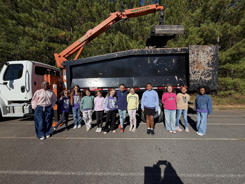 Students pose next to a garbage truck on a road lined with trees. One adult is wearing a pink jacket.