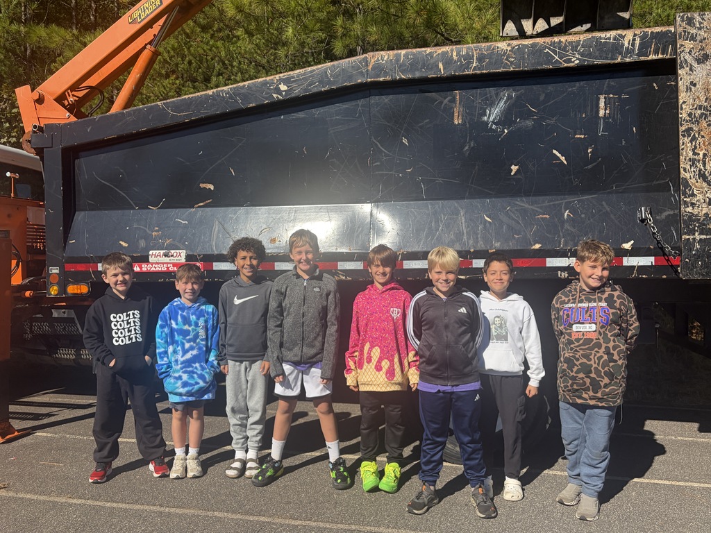 Several children in warm clothing stand in front of a large black dumpster on a paved surface.